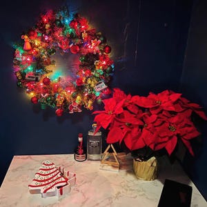 May include: A festive Christmas wreath with colorful lights and ornaments hangs on a dark blue wall. A poinsettia plant with vibrant red leaves sits next to a table with a Christmas tree-shaped cookie and bottles.