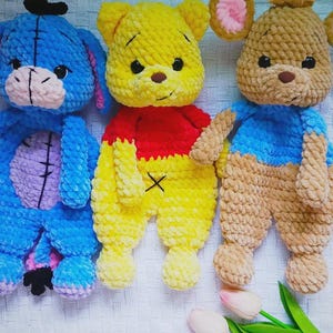May include: Three handmade crochet plush toys featuring a blue donkey with a pink muzzle, a yellow bear wearing a red shirt, and a tan mouse with pink inner ears. Each character is crafted from soft, chunky yarn with dark safety eyes and stitched details.