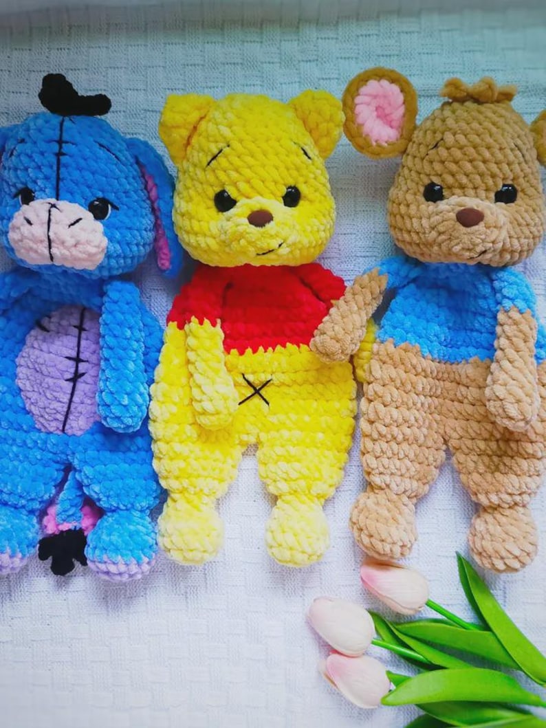 May include: Three handmade crochet plush toys featuring a blue donkey with a pink muzzle, a yellow bear wearing a red shirt, and a tan mouse with pink inner ears. Each character is crafted from soft, chunky yarn with dark safety eyes and stitched details.