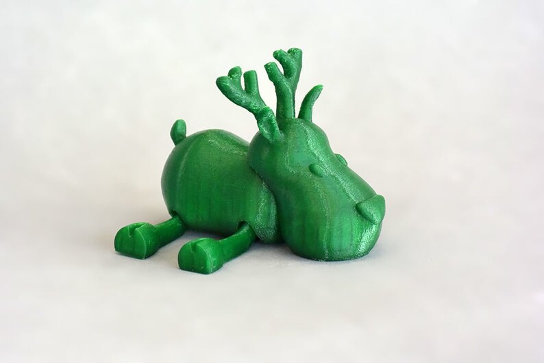 May include: A green, 3D-printed toy deer figurine. The deer is lying down with its legs extended and has antlers. The toy is a solid green color and appears to be made of plastic. The background is plain white.