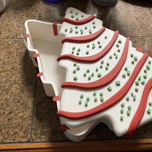 May include: A white, Christmas tree-shaped container with a lid. The tree design features red stripes and small green square decorations. The container is open, revealing its interior, and sits on a brown surface.