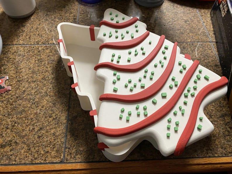 May include: A white, Christmas tree-shaped container with a lid. The tree design features red stripes and small green square decorations. The container is open, revealing its interior, and sits on a brown surface.