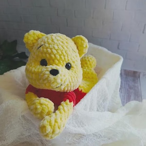 May include: A handmade crochet plush toy shaped like a bear with yellow yarn fur, a red shirt, and black plastic eyes. The bear has rounded ears, a small black nose, and a stitched mouth, crafted from soft, textured yarn in a seated position.