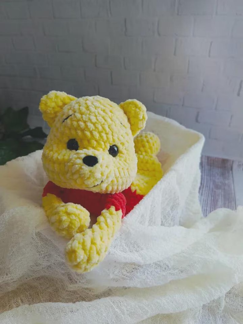 May include: A handmade crochet plush toy shaped like a bear with yellow yarn fur, a red shirt, and black plastic eyes. The bear has rounded ears, a small black nose, and a stitched mouth, crafted from soft, textured yarn in a seated position.