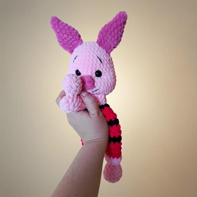 May include: A crocheted plush toy resembling a small pink pig character with long dark pink ears, black eyes, and a dark pink nose. The body features a striped pattern of alternating bright pink and black sections, crafted from soft yarn.