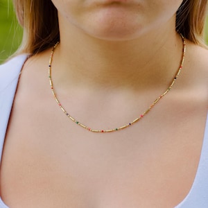 May include: A delicate gold chain necklace featuring a repeating pattern of small, cylindrical gold beads interspersed with tiny, vibrant multi-colored seed beads in shades of red, green, blue, and orange, resting against the base of the neck.