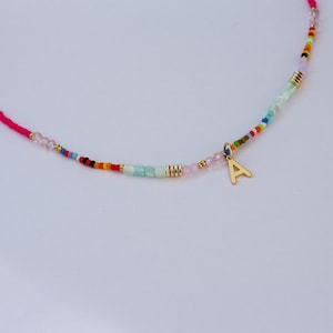 May include: A colorful beaded necklace with a gold-colored letter "A" pendant. The necklace features a pink cord and a variety of small, colorful beads, including pink, blue, green, and gold. The beads are arranged in a pattern.