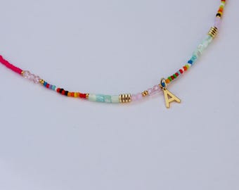 May include: A colorful beaded necklace with a gold-colored letter "A" pendant. The necklace features a pink cord and a variety of small, colorful beads, including pink, blue, green, and gold. The beads are arranged in a pattern.