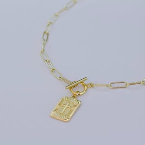 May include: Gold-toned necklace featuring a rectangular pendant with a cross design. The pendant is attached to a chain with an elongated link design and a toggle clasp. The necklace is set against a soft, neutral background.