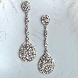 May include: A pair of silver dangle earrings with a teardrop-shaped design. Each earring features a delicate chain with small clear stones and a larger teardrop-shaped pendant with a floral pattern.