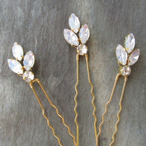 May include: Three gold hair pins with clear crystal embellishments in the shape of leaves. The pins are wavy and have a delicate design.