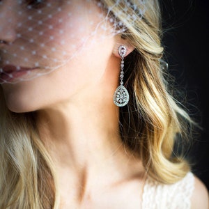 Pu&ograve; includere: Una donna indossa una camicetta bianca in pizzo con un velo e lunghi capelli biondi. Indossa lunghi orecchini a forma di lacrima con dettagli in cristallo.