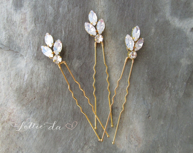 May include: Three gold hair pins with clear crystal embellishments in the shape of leaves. The pins are wavy and have a delicate design.