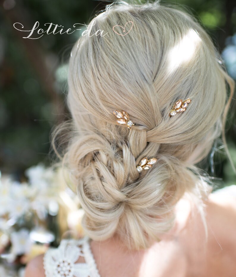 May include: A woman with blonde hair wearing a white dress with a lace pattern. Her hair is styled in a bun with three gold hair pins with clear rhinestones. The hair pins are shaped like leaves.
