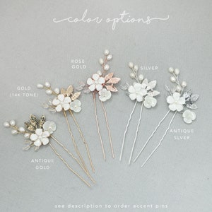 May include: Six hair pins with white pearl flowers and leaves in different metal finishes: antique gold, gold (14k tone), rose gold, silver, and antique silver. The text "color options" is written above the pins.