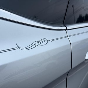 May include: Silver car door with a decorative gray vinyl decal. The decal features a stylized design with curved lines and a central swirl. Two parallel gray lines run horizontally along the car's side.