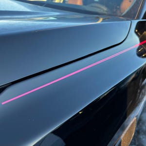 May include: A close-up shot of a black car's bodywork, showcasing a bright pink stripe. The car's surface is highly polished, reflecting the environment. The image focuses on the car's design and the contrast between the black and pink.