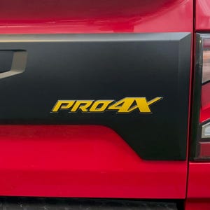 May include: Close-up of a red truck bed with a black tailgate panel. The panel features the yellow text "PRO4X". The truck's taillight is visible on the right side of the image.