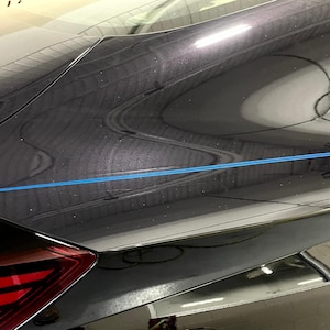 Puede incluir: Una sección de un coche gris oscuro con una raya azul. La imagen muestra la parte trasera del coche, incluyendo un piloto trasero rojo y el panel curvo de la carrocería. La superficie del coche refleja el entorno.