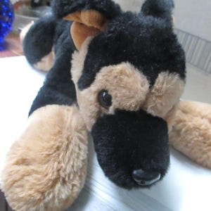 May include: A plush German Shepherd dog toy with black and tan fur. The toy has a black nose, brown eyes, and tan paws. The dog is lying down on a white surface.