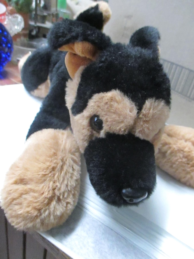 May include: A plush German Shepherd dog toy with black and tan fur. The toy has a black nose, brown eyes, and tan paws. The dog is lying down on a white surface.