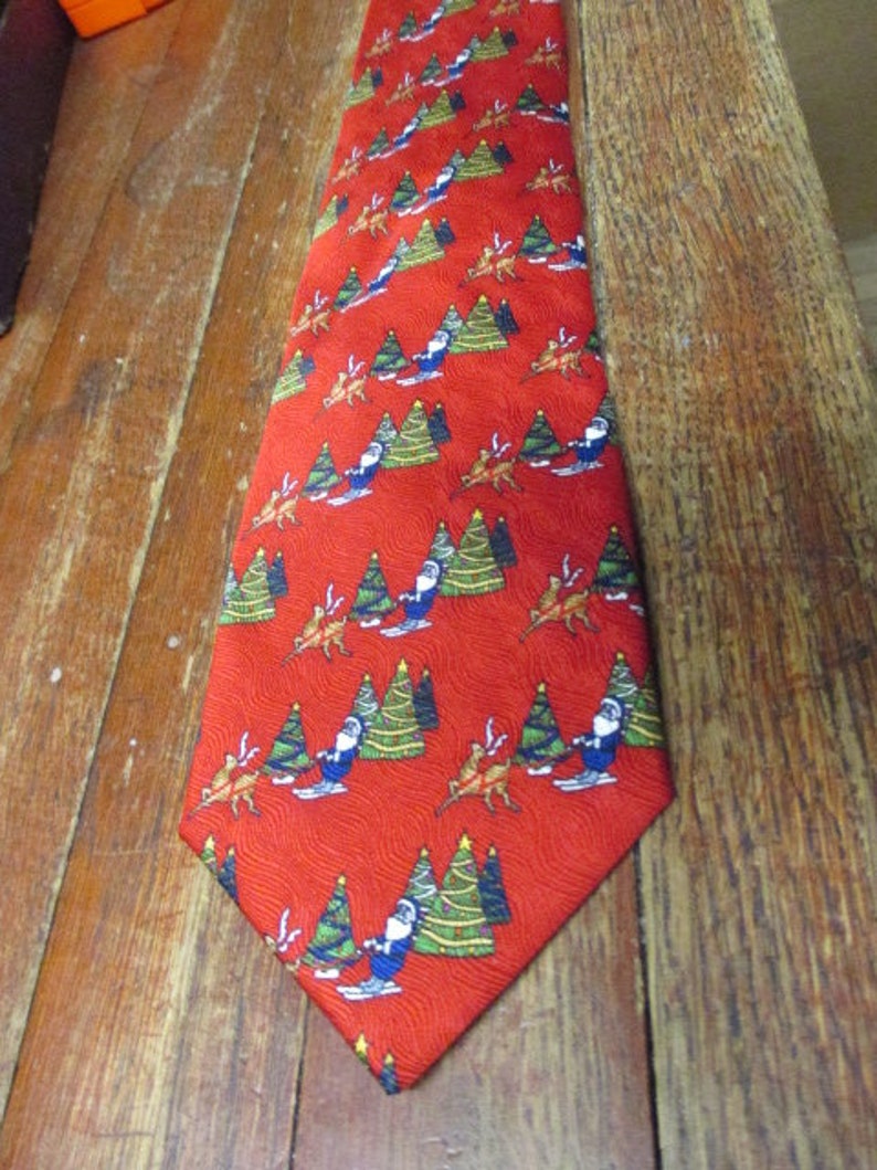 May include: Red Christmas tie with a repeating pattern of Santa Claus skiing, Christmas trees, and reindeer. The tie is made of a shiny fabric and is suitable for the holiday season.