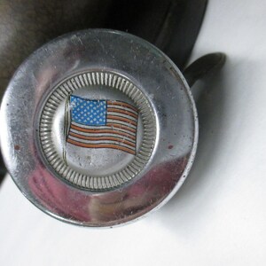 May include: A circular silver-toned metal object with a polished finish and a central inset featuring a graphic of the American flag. The flag design displays a blue field with white stars and red and white stripes, surrounded by a ridged border.