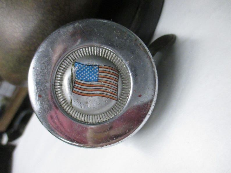 May include: A circular silver-toned metal object with a polished finish and a central inset featuring a graphic of the American flag. The flag design displays a blue field with white stars and red and white stripes, surrounded by a ridged border.