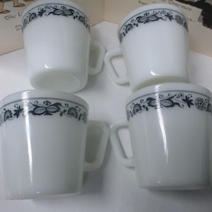 May include: Four white mugs with a dark blue floral design around the rim. Each mug has a handle and a slightly flared shape. The mugs are arranged in a square, showcasing their clean, classic design.