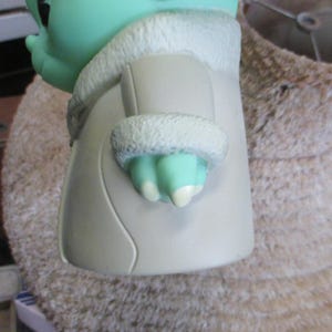 May include: A green figurine of a character with large ears and a tan coat with a white, furry trim. The figurine has a light green hand with three fingers and white nails. The character's face is visible, with a black eye and a small nose.