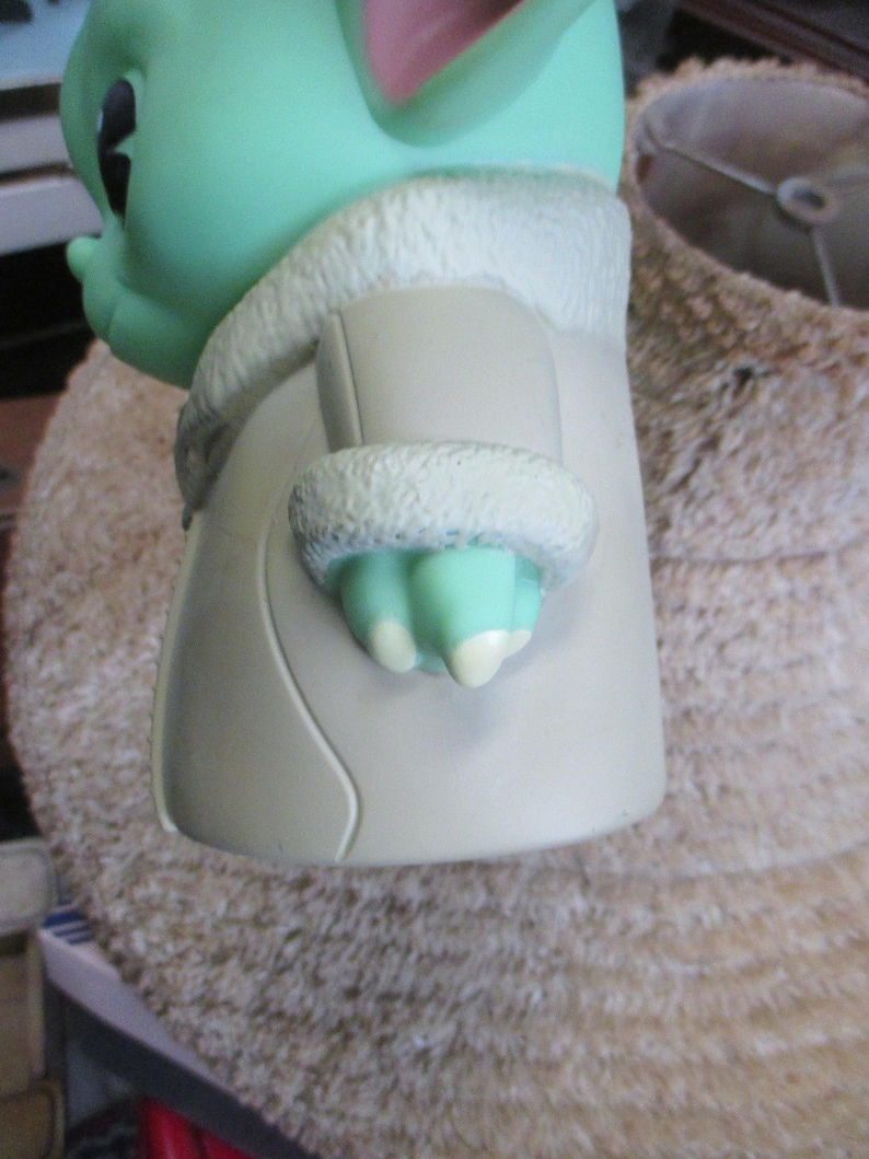 May include: A green figurine of a character with large ears and a tan coat with a white, furry trim. The figurine has a light green hand with three fingers and white nails. The character's face is visible, with a black eye and a small nose.