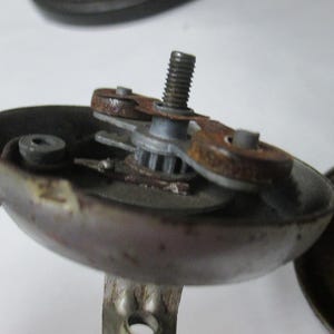 May include: A disassembled metal bicycle bell mechanism featuring a central threaded bolt, a small gear, and two circular components with visible rust. The bell housing is grey with signs of wear, and the internal striking assembly is exposed.
