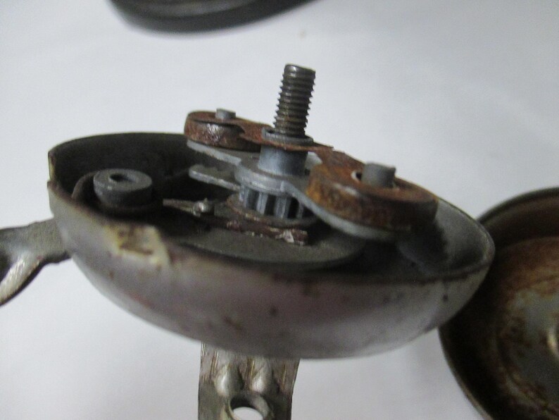 May include: A disassembled metal bicycle bell mechanism featuring a central threaded bolt, a small gear, and two circular components with visible rust. The bell housing is grey with signs of wear, and the internal striking assembly is exposed.