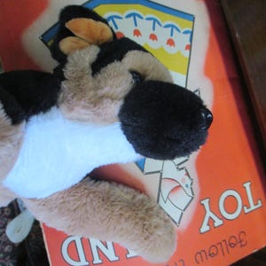 May include: A plush German Shepherd dog toy with black, tan, and white fur, lying on a vintage book cover. The book cover is orange with the words "TOY LAND" and "Follow the Dots" printed on it.