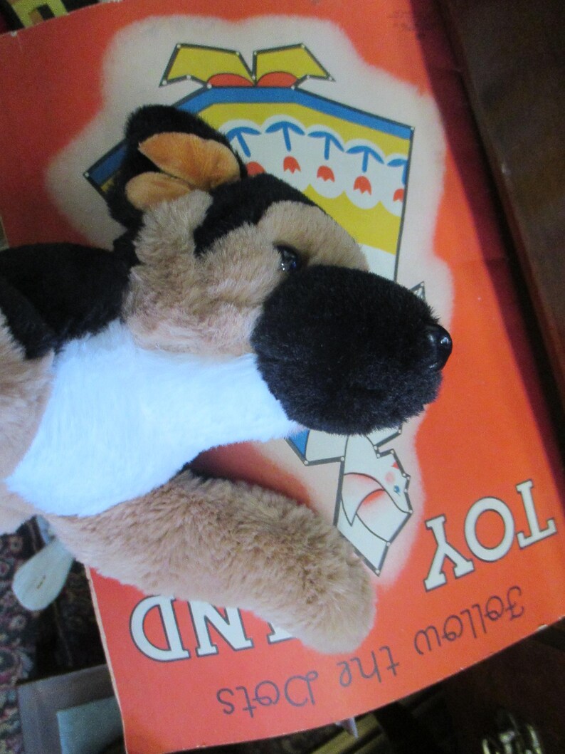 May include: A plush German Shepherd dog toy with black, tan, and white fur, lying on a vintage book cover. The book cover is orange with the words "TOY LAND" and "Follow the Dots" printed on it.