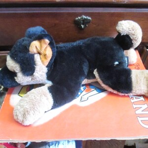 May include: A plush toy dog in black and tan, lying on an orange book. The dog has floppy ears and a soft, cuddly appearance. The book has the word "LAND" visible on the cover. The toy is a classic children's item.