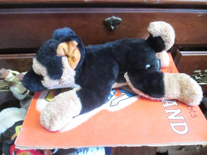 May include: A plush toy dog in black and tan, lying on an orange book. The dog has floppy ears and a soft, cuddly appearance. The book has the word "LAND" visible on the cover. The toy is a classic children's item.