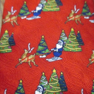May include: Red Christmas tie with a repeating pattern of Santa Claus skiing, reindeer, and decorated Christmas trees. The trees are green with yellow stars, and the reindeer are brown. The Santa Claus figures are wearing blue suits.