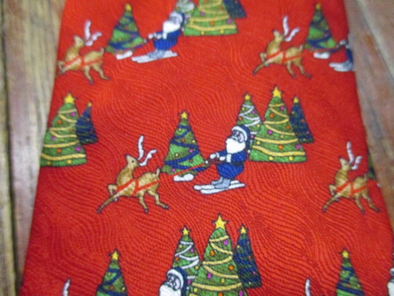 May include: Red Christmas tie with a repeating pattern of Santa Claus skiing, reindeer, and decorated Christmas trees. The trees are green with yellow stars, and the reindeer are brown. The Santa Claus figures are wearing blue suits.