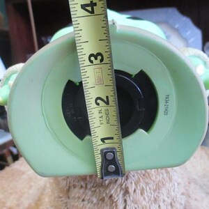 May include: A light green ceramic piggy bank shaped like a character with large ears. The opening is approximately 2 inches wide. The bank is being measured with a yellow tape measure.