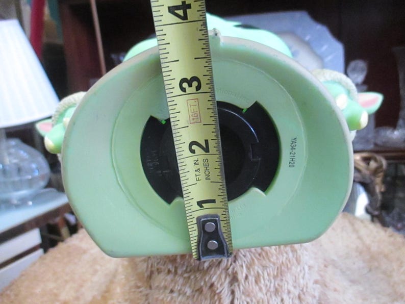 May include: A light green ceramic piggy bank shaped like a character with large ears. The opening is approximately 2 inches wide. The bank is being measured with a yellow tape measure.