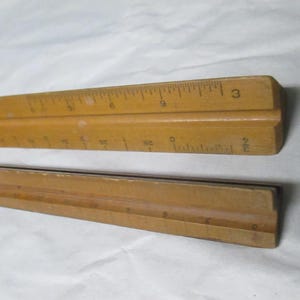 May include: Two wooden rulers, one with measurements in inches. The rulers are light brown and appear to be vintage. The top ruler shows measurements up to 3 inches.