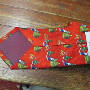 May include: Red necktie with a repeating pattern of green Christmas trees and figures. The tie has a burgundy backing and a tag that reads "Cape Cod Neckwear".