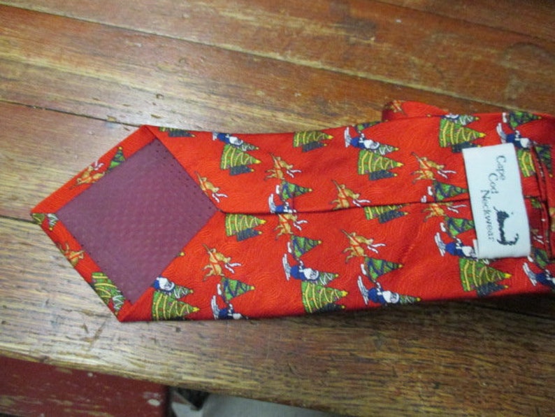 May include: Red necktie with a repeating pattern of green Christmas trees and figures. The tie has a burgundy backing and a tag that reads "Cape Cod Neckwear".