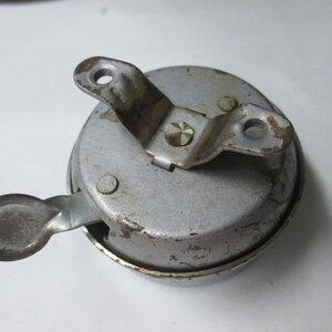 May include: A circular silver-toned metal bicycle bell with a weathered finish. The bell features a top-mounted bracket with two screw holes and a side-mounted thumb lever for activation. The surface shows signs of wear, oxidation, and minor rust.