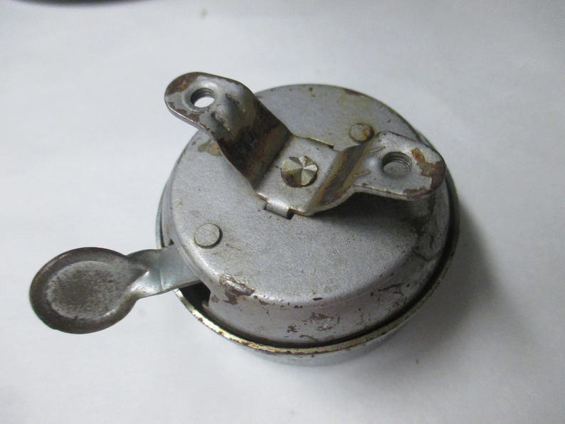 May include: A circular silver-toned metal bicycle bell with a weathered finish. The bell features a top-mounted bracket with two screw holes and a side-mounted thumb lever for activation. The surface shows signs of wear, oxidation, and minor rust.