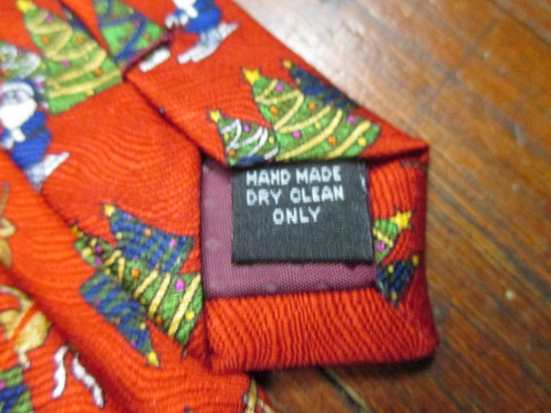 May include: Red Christmas tie with a repeating pattern of Christmas trees and Santa Claus figures. The tie has a label that reads "HAND MADE DRY CLEAN ONLY".