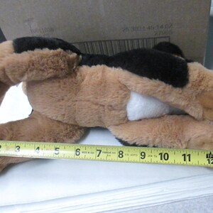 May include: A plush dog toy, primarily brown with black patches and a white belly. The toy is measured with a yellow tape measure, approximately 12 inches long.