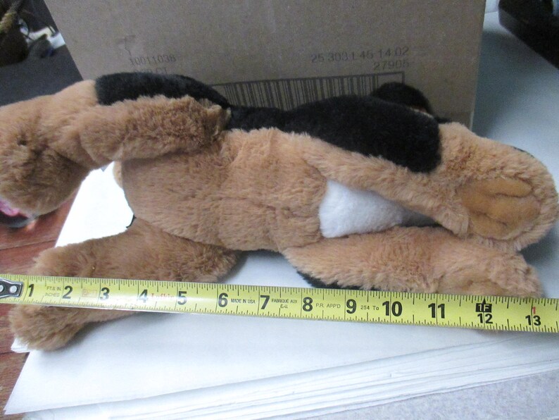 May include: A plush dog toy, primarily brown with black patches and a white belly. The toy is measured with a yellow tape measure, approximately 12 inches long.