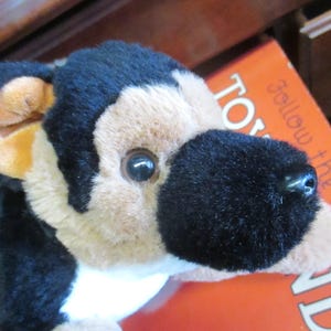 May include: A plush German Shepherd dog toy with black, tan, and cream-colored fur. The toy has a black nose and a detailed face. The toy is resting on an orange book with the words "Follow the Dots" visible.